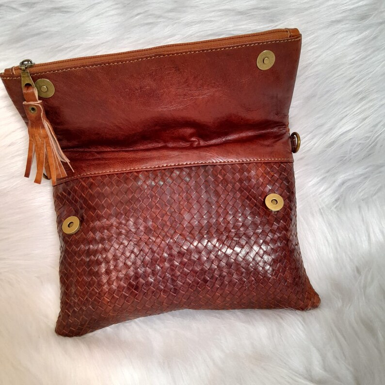 May include: A brown leather clutch with a woven pattern on the front. The clutch has a zipper closure and a tassel. The top flap is secured with two magnetic snaps. The bag is open, revealing the interior.