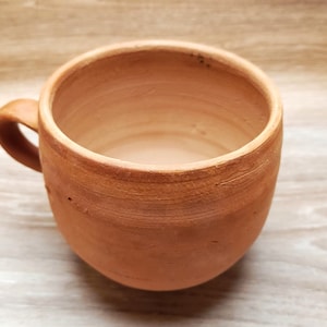 Peut inclure: Une tasse en céramique faite à la main, de couleur terre cuite, avec un corps arrondi et une petite anse incurvée. La tasse a un aspect rustique et terreux, avec une texture visible et de légères variations de couleur. L'intérieur est lisse.