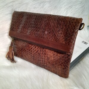 May include: A brown leather clutch with a woven pattern. The clutch has a zipper closure, a leather strap, and a tassel. The bag is rectangular and appears to be made of genuine leather.