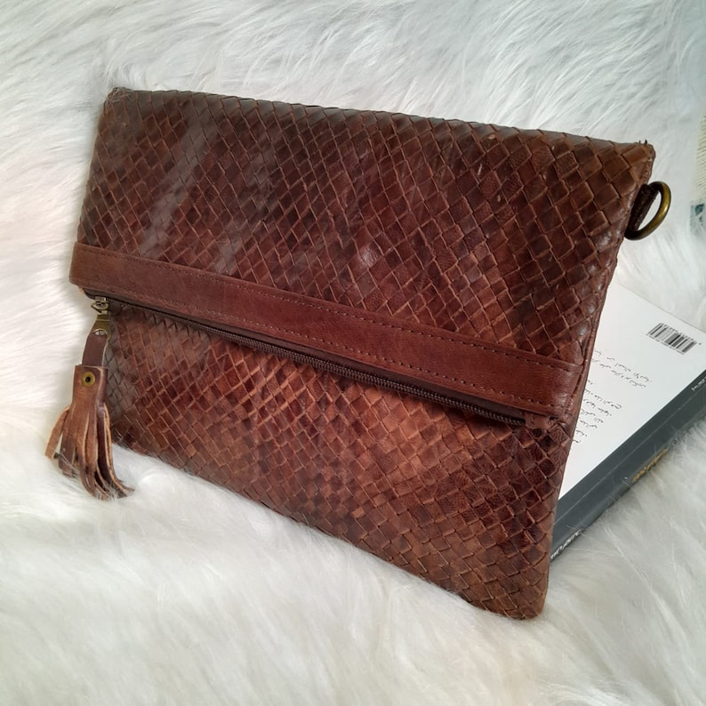 May include: A brown leather clutch with a woven pattern. The clutch has a zipper closure, a leather strap, and a tassel. The bag is rectangular and appears to be made of genuine leather.