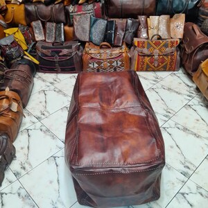 May include: A large, brown leather bag with white stitching is the focal point, surrounded by a variety of leather bags and satchels in various colors and designs. The bags are displayed on a tiled floor.