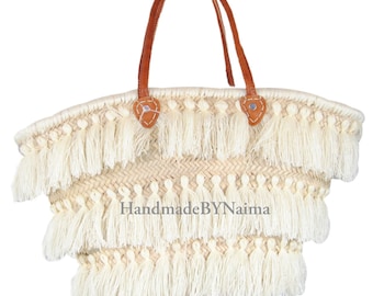 Może przedstawiać: Tkana torba typu tote ze słomy z kremowymi frędzlami i brązowymi skórzanymi uchwytami. Torba zawiera napis "HandmadeBYNaima" i jest zaprojektowana z wieloma warstwami frędzli.