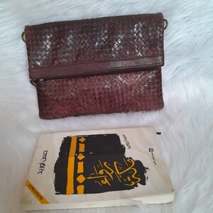 May include: A dark brown, woven leather clutch with a zipper closure and a small handle. A book with Arabic text and a black and yellow design is below the clutch.