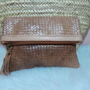 May include: A tan leather clutch with a woven pattern. The clutch has a zipper closure and a tassel. The top of the clutch is a lighter tan color. The clutch is folded over and appears to be empty.