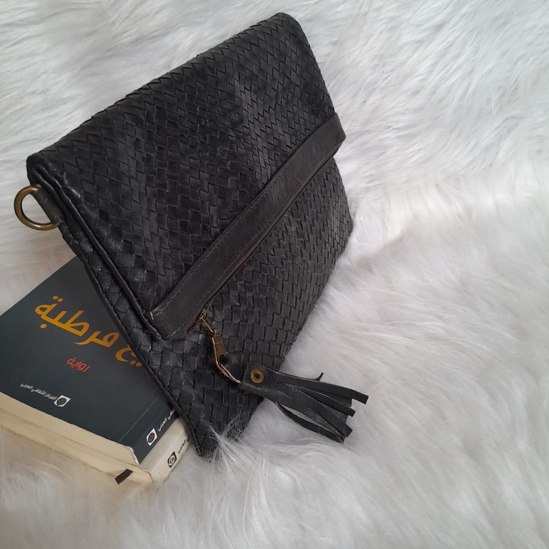 May include: A dark gray woven leather clutch with a fold-over flap. The clutch features a zipper closure with a tassel and a gold-colored ring. A book with Arabic text is partially visible.