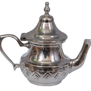 Peut inclure: Une théière argentée avec un motif décoratif. La théière a une poignée incurvée, un bec verseur et un couvercle avec un bouton. Le corps de la théière présente un motif en relief. La théière est probablement en métal.
