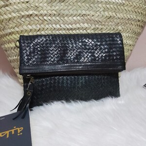 May include: Black woven leather clutch with a fold-over flap and a zipper closure. The clutch has a wrist strap and a tag attached. The background includes a woven basket and white fur.