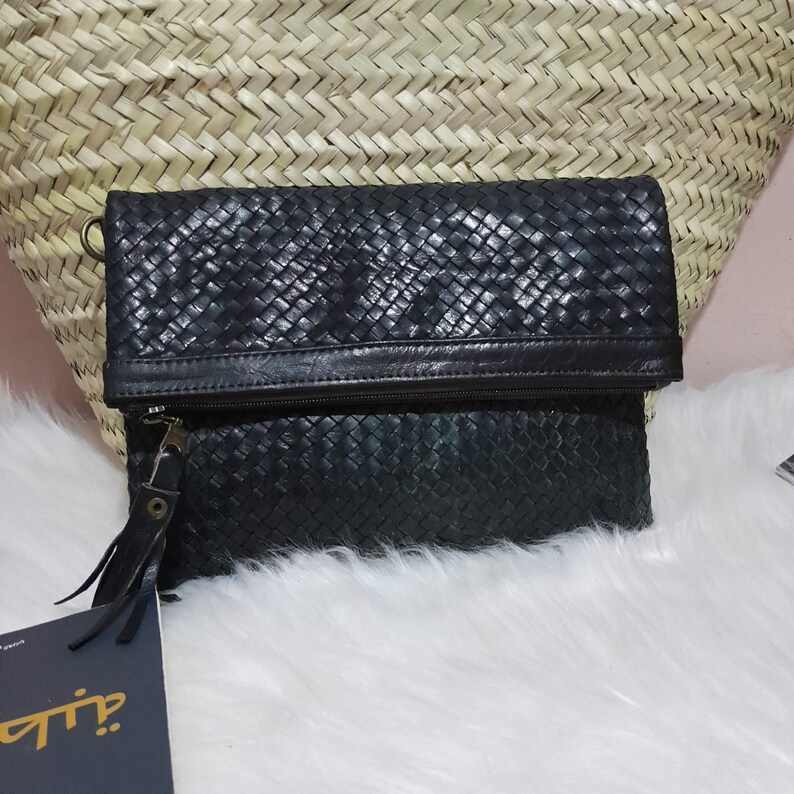 May include: Black woven leather clutch with a fold-over flap and a zipper closure. The clutch has a wrist strap and a tag attached. The background includes a woven basket and white fur.