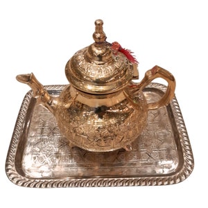 Peut inclure: Une théière dorée avec des motifs en relief complexes, un couvercle décoratif et une poignée incurvée, posée sur un plateau argenté avec une bordure détaillée. La théière a un petit pompon rouge attaché au couvercle.