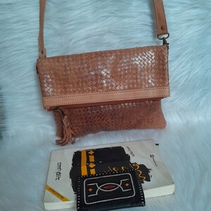 May include: A light brown leather crossbody bag with a woven pattern. The bag has a long strap, a zipper closure, and a tassel. A book and a small black wallet with white and red stitching are also visible.
