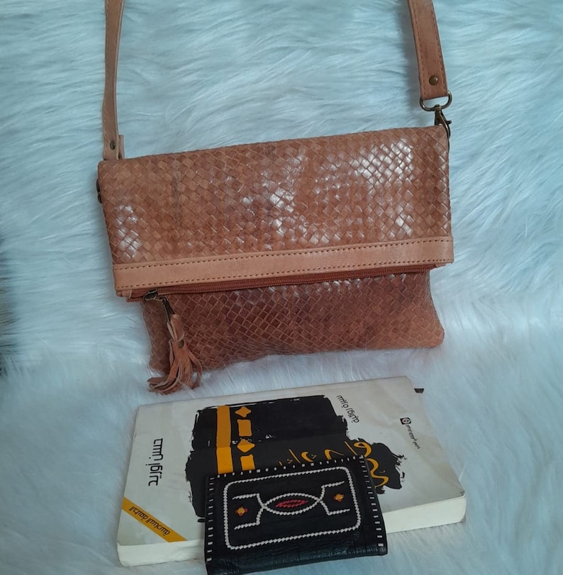 May include: A light brown leather crossbody bag with a woven pattern. The bag has a long strap, a zipper closure, and a tassel. A book and a small black wallet with white and red stitching are also visible.