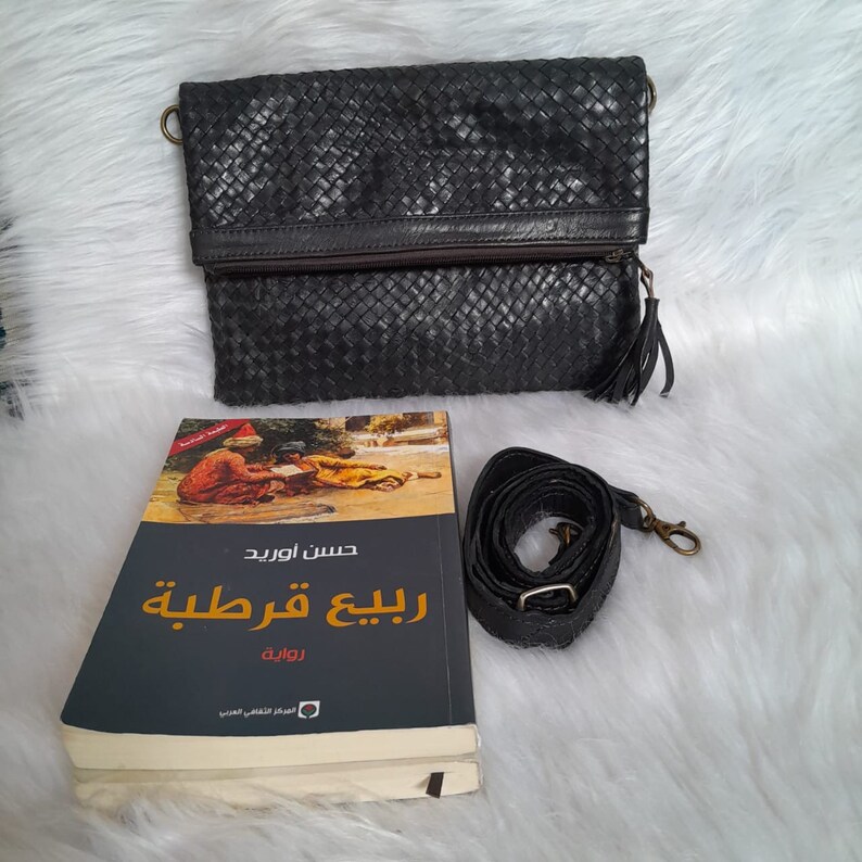 May include: Black woven leather clutch with a tassel zipper pull and a detachable shoulder strap. A book with Arabic text and a colorful illustration is next to the clutch.