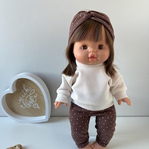 May include: A brown doll with brown hair wearing a white sweater and brown polka dot leggings. The doll is wearing a brown headband with white polka dots. The doll is standing on a white surface. The doll is looking at the camera.