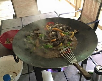 Puede incluir: Una sartén grande, redonda y de hierro fundido negro se encuentra sobre un soporte de metal negro con una llama azul debajo. La sartén está llena de una mezcla de pimientos, cebollas y carne cortados en rodajas. Una espátula descansa en el borde de la sartén. Un rollo de papel de cocina está sobre la mesa junto a la sartén.