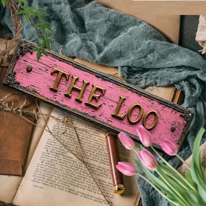 May include: A rectangular sign with a distressed pink finish and a dark, ornate border. The sign features raised, metallic gold lettering that spells out THE LOO. The piece is accented with small, dark decorative corner details.