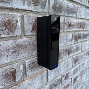 May include: A black rectangular security camera is mounted on a weathered brick wall. The camera has a glossy front panel, a small lens, and a blue light at the bottom. The brick wall is painted white, with red and brown tones showing through.