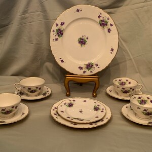 May include: A set of vintage teacups and saucers with a delicate floral pattern featuring purple violets and green leaves. The set includes a larger plate with the same floral design, displayed on a gold stand.