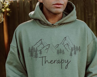 Può includere: Una felpa con cappuccio verde con una grafica nera di una catena montuosa e alberi e il testo "Therapy" stampato sul davanti.