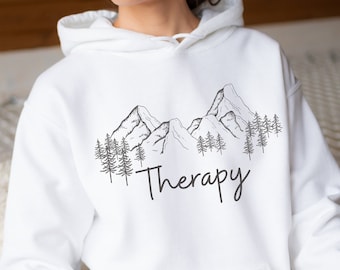 Può includere: Felpa con cappuccio bianca con una grafica in bianco e nero di montagne e alberi. Il testo "Therapy" è stampato sotto la grafica.