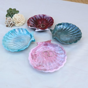 May include: Four seashell-shaped trinket dishes in shades of blue, red, green, and pink. Each dish has a textured surface and holds a gold ring. Decorative flowers are in the upper left corner. These dishes are ideal for holding jewelry.