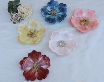 Könnte beinhalten: Sechs blütenförmige Ringschalen in verschiedenen Farben, darunter Blau, Rosa, Gelb und Rot. Jede Schale hat eine glitzernde Mitte, und einige halten Ringe. Dekorative Sukkulenten und Holzblumen sind ebenfalls vorhanden.