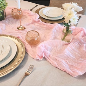 May include: A pink table runner with a gold charger plate, a white dinner plate, and a white plate with a silver design. A clear glass vase with white flowers sits on the table runner.