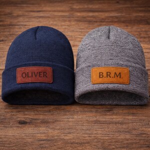Puede incluir: Un gorro de punto azul marino con un parche de cuero marrón rectangular grabado con el nombre OLIVER y un gorro de punto gris jaspeado con un parche de cuero tostado rectangular grabado con las iniciales B.R.M. Ambos gorros tienen un borde doblado.
