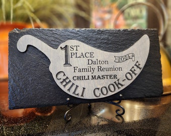 Pode incluir: Uma placa retangular de ardósia preta com um recorte de madeira clara em forma de pimenta. A madeira está gravada com o texto 1ST PLACE, Dalton Family Reunion, 2024, CHILI MASTER e CHILI COOK-OFF. A placa repousa sobre um suporte de metal preto.