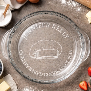 May include: A clear glass pie dish with handles, featuring the text "JENNIFER KELLY QUALITY BAKED GOODS SINCE 2018" and a chef's hat design. The dish is round and appears to be new.