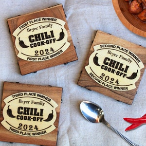 May include: Three wooden plaques with a yellow background and black text. The plaques are for a chili cook-off and say "First Place Winner", "Second Place Winner", and "Third Place Winner". Each plaque has the name "Bryer Family" and the year "2024".
