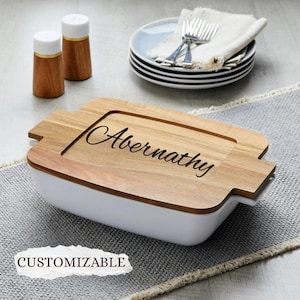 May include: A white rectangular baking dish with a wooden lid engraved with the name "Abernathy." Two wooden salt shakers and a stack of plates with silverware are in the background. The word "CUSTOMIZABLE" is in the lower left corner.