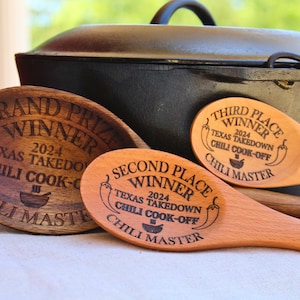 May include: Three wooden spoons with engraved text, each indicating a different place in a chili cook-off. The spoons are arranged on a white surface with a black cast iron pot in the background. The text on the spoons reads "Grand Prize Winner", "Second Place Winner", and "Third Place Winner".