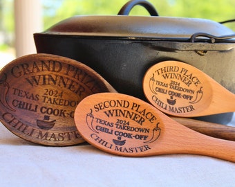 以下が含まれることがあります： Grand Prize Winner、Second Place Winner、Third Place Winnerと刻印された3本の木製スプーン。2024 Texas Takedown Chili Cook-Off Chili Masterの文字と唐辛子のイラストが描かれています。スプーンは黒い鋳鉄製の鍋の前に置かれています。