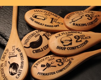 May include: Wooden spoons with engraved text and illustrations. The spoons are engraved with text such as "Mason Family 2024 The Great Bake-Off 1st Place" and "Madison Group 2024 Cast Iron Cook-Off 1st Place". The spoons are for cooking competitions.