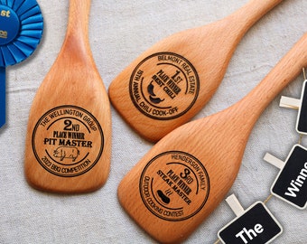 May include: Three light brown wooden cooking spoons with custom laser-engraved text and graphics. One spoon reads 2nd Place Winner Pit Master, another 1st Place Winner Best Chili, and the third 3rd Place Winner Steak Master. A blue 1st Place ribbon is included.