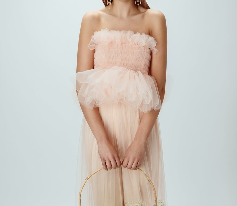 May include: A woman wearing a pink tulle dress with a ruffled bodice. She is holding a wicker basket with white flowers.