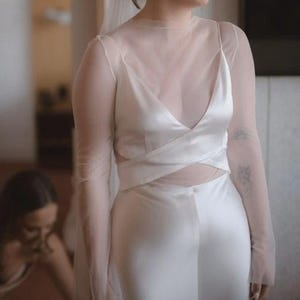 May include: A white bridal ensemble featuring a satin top with a crisscross design and a sheer long-sleeved overlay. The outfit includes matching pants. The bride is wearing a veil.