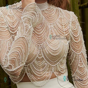 May include: A long-sleeved, high-neck top featuring intricate white pearl and clear sequin embellishments arranged in scalloped, wave-like patterns across a sheer, nude-toned base. The garment includes the text Label Wardrobe printed on it.
