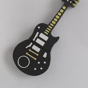Könnte beinhalten: Ein schwarzer Gitarrenmagnet mit weißen und gelben Akzenten. Der Korpus hat einen weißen Kreis und Punkte, mit weißen Rechtecken und gelben Quadraten entlang des Halses. Die Kopfplatte hat gelbe Details.