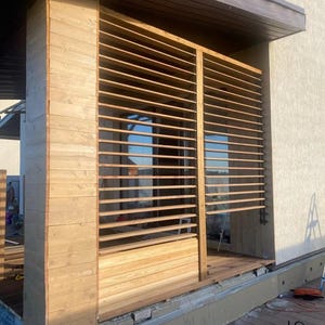 Puede incluir: Estructura exterior de madera con lamas horizontales, posiblemente una pantalla de privacidad o un elemento decorativo. Construida con madera clara, está fijada a un edificio con un tejado marrón. El diseño permite la entrada de luz filtrada y ventilación.
