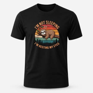May include: A black short-sleeved t-shirt featuring a graphic of a brown sloth resting on a tree branch. The design includes a sunset background with silhouettes of pine trees and the text I'M NOT SLEEPING I'M RESTING MY EYES in orange.