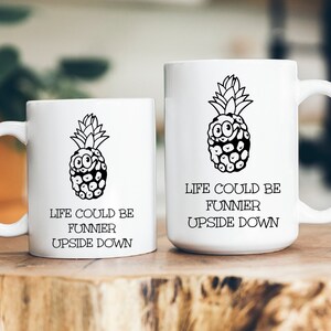 May include: Two white ceramic mugs with black pineapple designs and the text "Life could be funnier upside down".