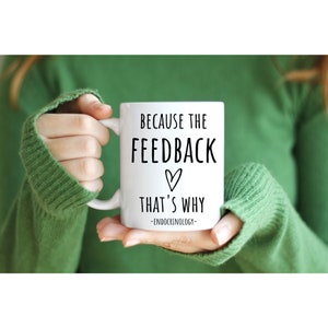May include: White ceramic mug with the text "BECAUSE THE FEEDBACK THAT'S WHY" in black, with a heart symbol, and the word "ENDOCRINOLOGY" below. The mug is held by a person wearing a green sweater.