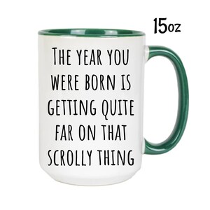May include: White ceramic mug with a green rim and handle. The mug has black text that reads "The year you were born is getting quite far on that scrolly thing".