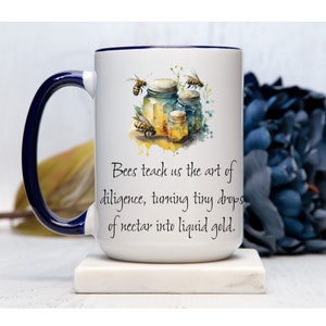 May include: White ceramic mug with a blue rim and handle. The mug features a watercolor illustration of honey jars and bees with the text "Bees teach us the art of diligence, turning tiny drops of nectar into liquid gold."