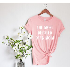 May include: Pink t-shirt with white text that reads "The Most Devoted OTTB Mom".
