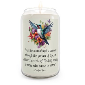 May include: A white candle with a glass jar features a hummingbird in flight surrounded by colorful flowers. The candle has a quote that reads "As the hummingbird dances through the garden of life, it whispers secrets of fleeting beauty to those who pause to listen." -Comfort Spice-