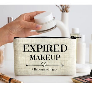 May include: A beige cosmetic bag with the text "EXPIRED MAKEUP" in black, with a heart and arrow design. A hand holds a white jar of cream above the bag. Other cosmetic products are in the background.