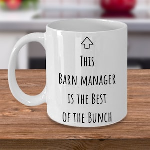 May include: White ceramic coffee mug with black text that reads "This Barn Manager is the Best of the Bunch". An arrow points to the text.