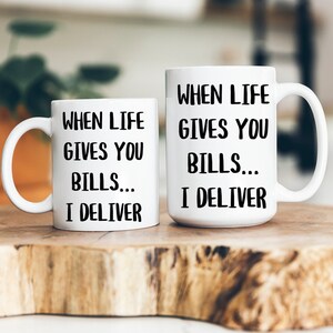 May include: Two white ceramic mugs with black text that reads "WHEN LIFE GIVES YOU BILLS... I DELIVER." The mugs are on a wooden surface. One mug is smaller than the other.